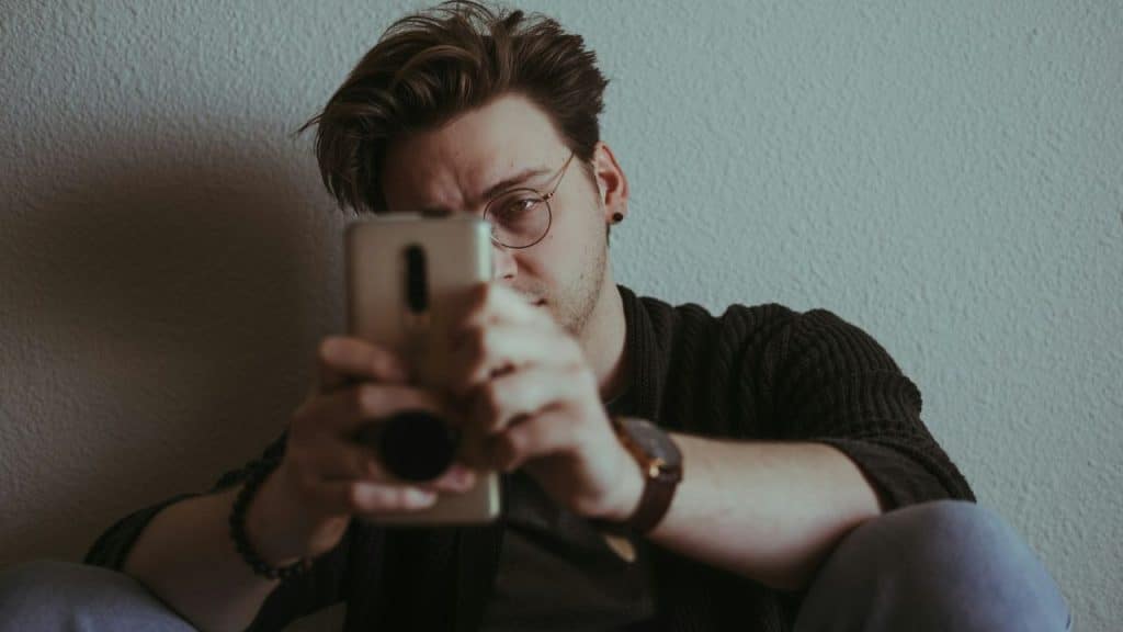 A man wearing glasses holds a smartphone in front of his face against a white wall.