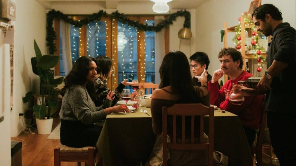 A group of people sits around a dinner table in a room decorated with string lights.