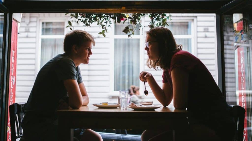 Two people sit at a small table inside a cafe, facing each other in profile.