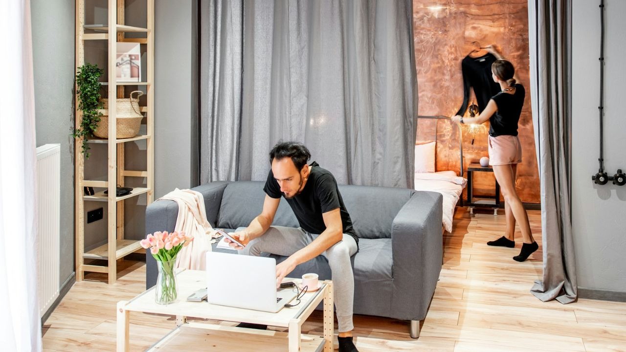 A man uses a laptop while a woman hangs a garment in a studio apartment.