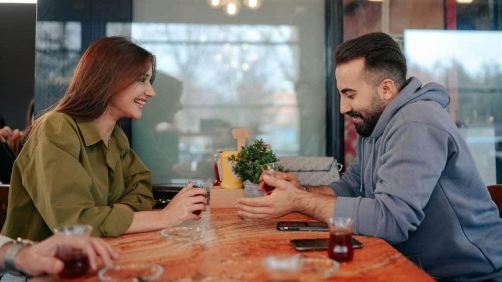 A man and woman sit at a wooden table holding small glasses of dark liquid.