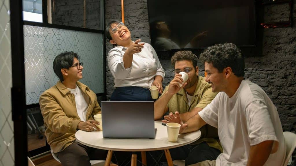 A group of four people laugh and talk around a table with a laptop.