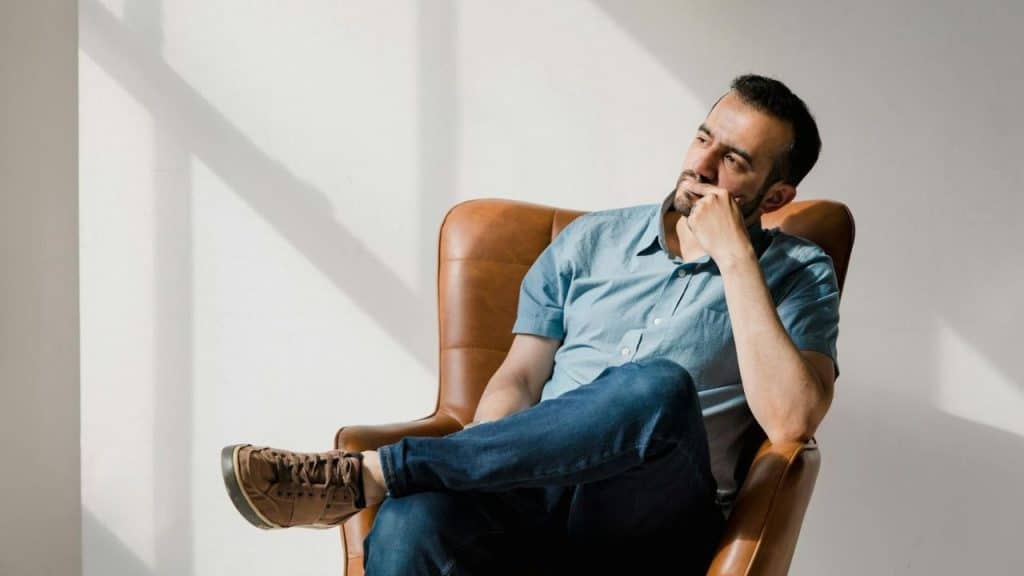 A man in a blue shirt and jeans sits in a tan leather armchair looking away.