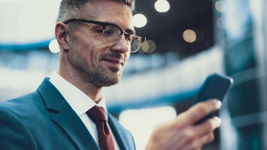 A man in a suit and glasses smiles while looking down at a smartphone in hand.