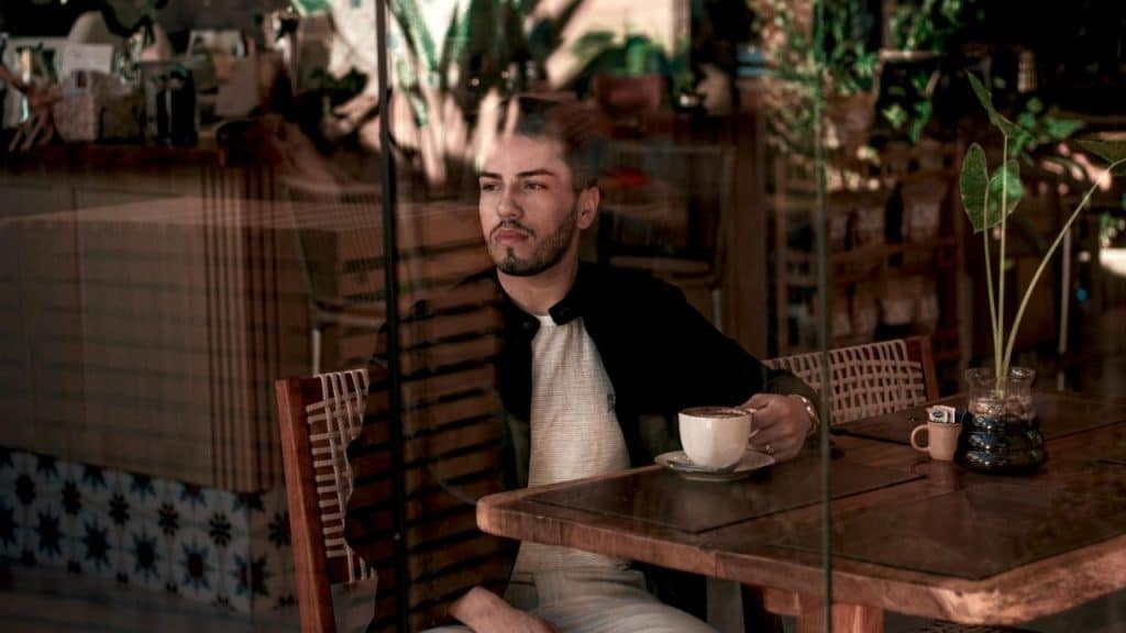 A man with a beard sits at a wooden cafe table holding a white coffee cup.