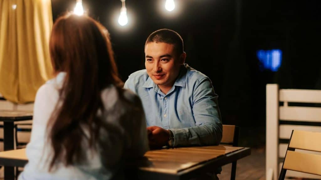 A man in a blue shirt sits at a table at night, looking at someone.