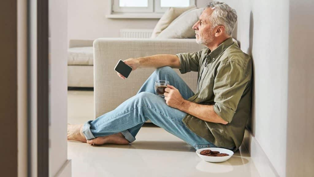 An older man sits on the floor holding a glass and a phone near cookies.