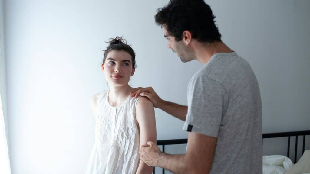 A man in a grey shirt places his hand on a woman's shoulder indoors.