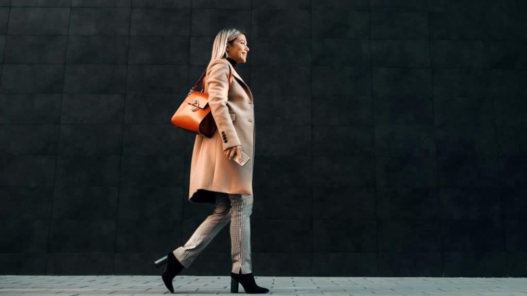 A smiling woman in a tan coat walks outdoors while carrying an orange bag.