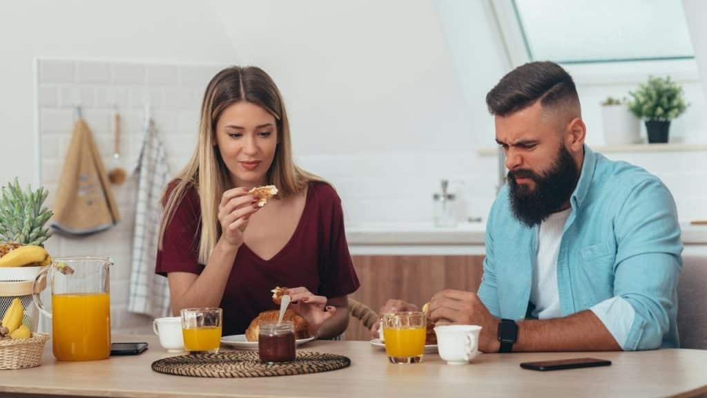 A woman eats a croissant while a man sitting beside her makes a frustrated expression.