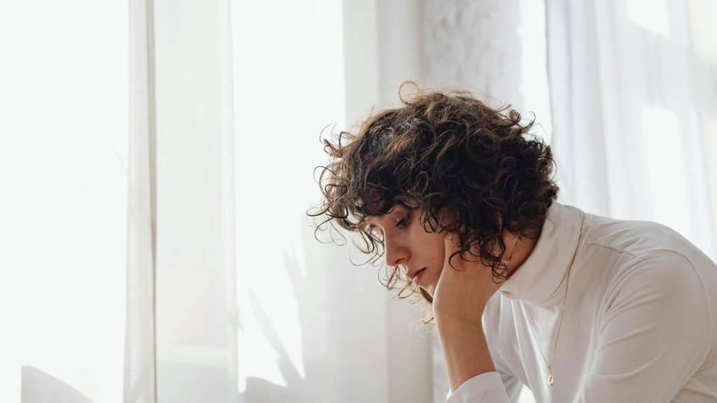A curly-haired person resting their head on a hand by a window.