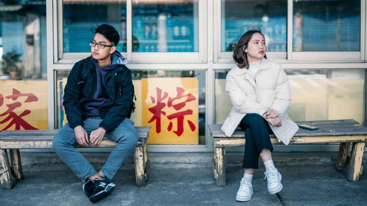 Two people sit on separate wooden benches outdoors, both looking away from each other.