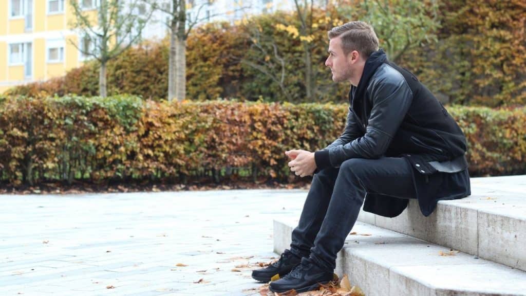 A man in a black jacket sits on stone steps outdoors looking off to the side.