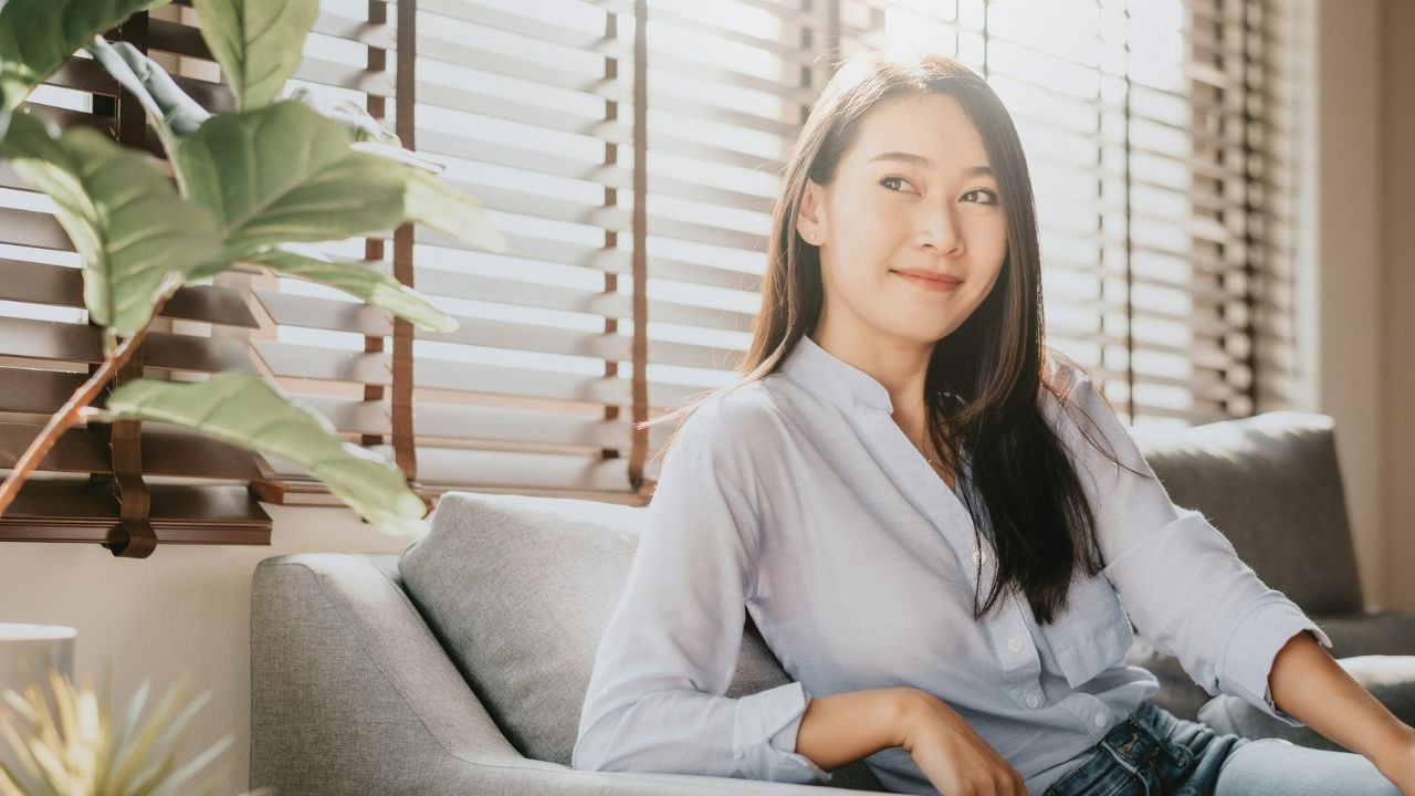 A smiling woman sits on a grey sofa near a window with wooden blinds.