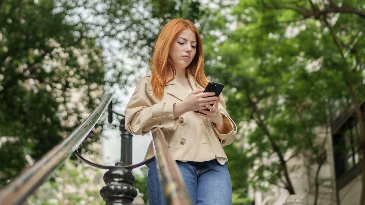 A woman with red hair stands on stairs outdoors while looking at her phone.