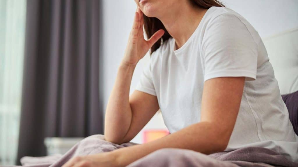 A woman sitting on a bed in a white shirt with her hand resting on her neck.