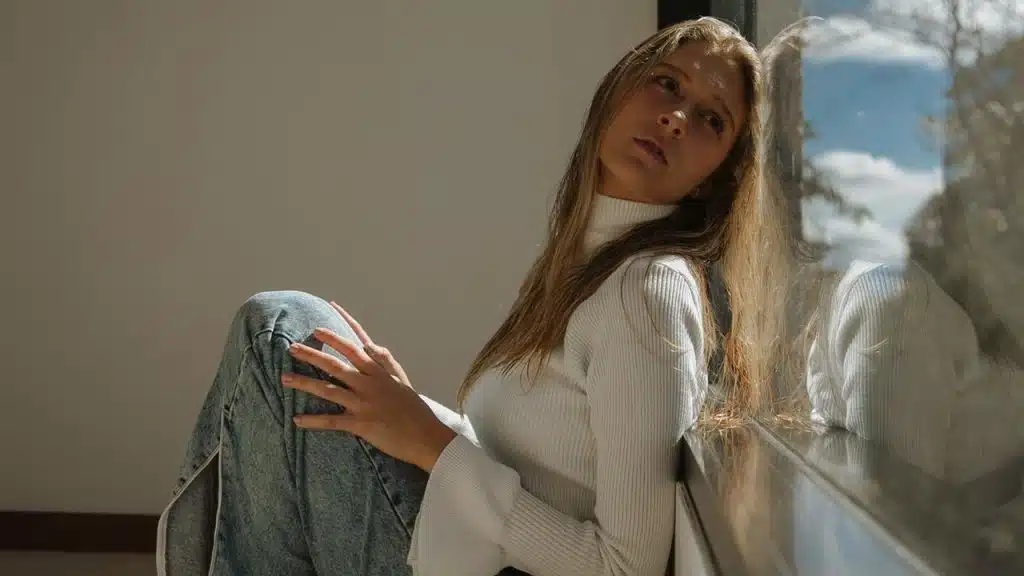 A woman in a white sweater sits by a sunlit window, gazing thoughtfully outside.