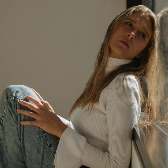 A woman in a white sweater sits by a sunlit window, gazing thoughtfully outside.