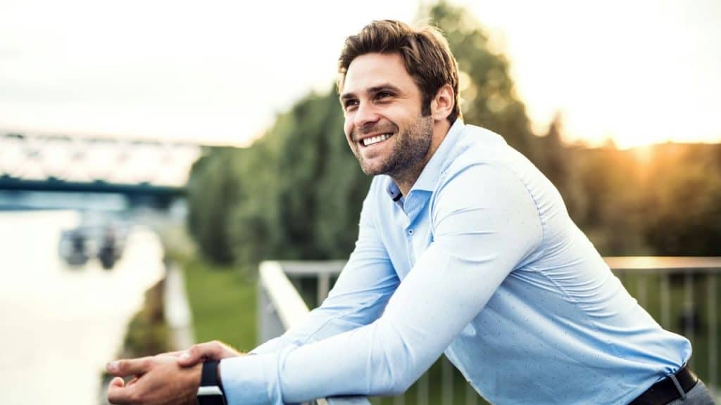 A smiling man in a blue shirt leans on a railing outdoors with trees in the background.