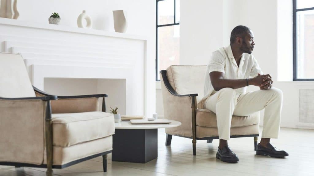A man in white clothing sits in a chair in a bright room with a white fireplace.