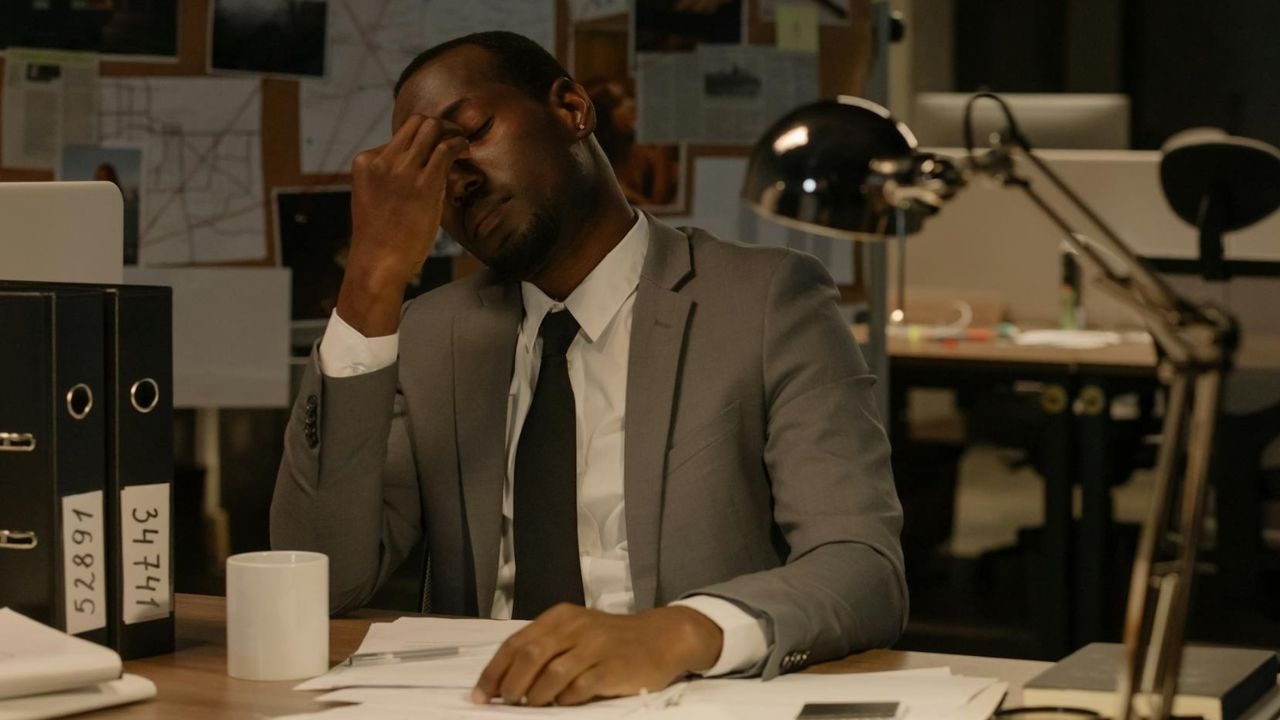 A man in a grey suit sits at a desk in an office while rubbing his eyes.