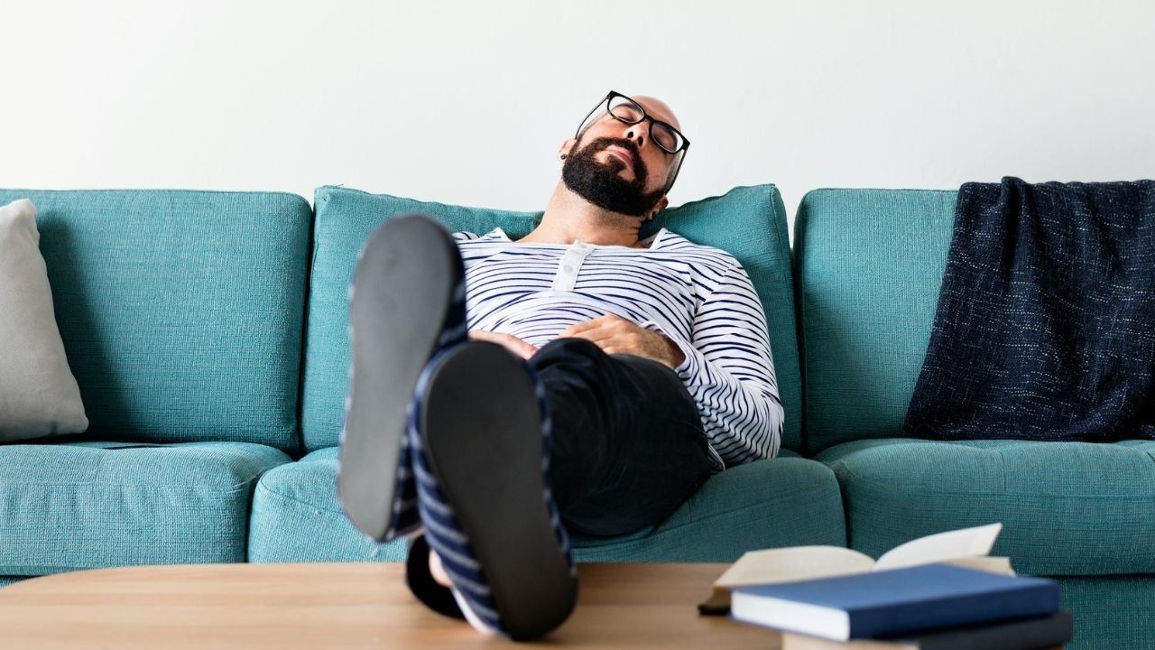 A bearded man in a striped shirt sleeps on a teal sofa with his feet up.