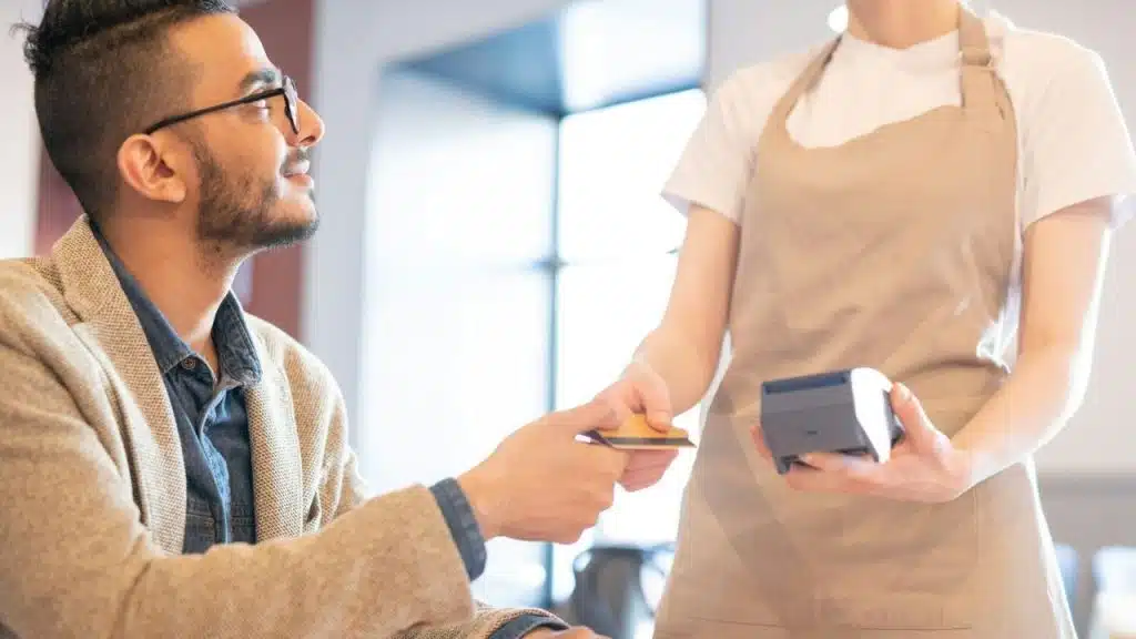 A seated man hands a credit card to a server holding a portable payment terminal.