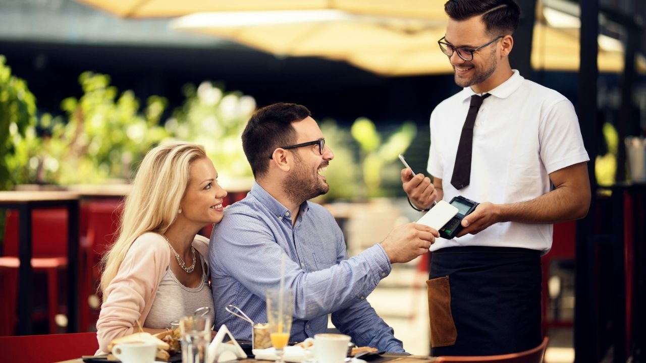A man at an outdoor table uses a smartphone to pay at a handheld terminal.