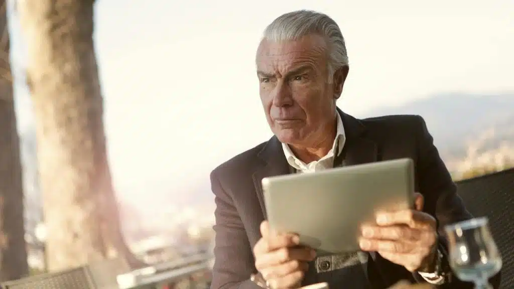 A man with grey hair holds a tablet while looking away with a focused expression.