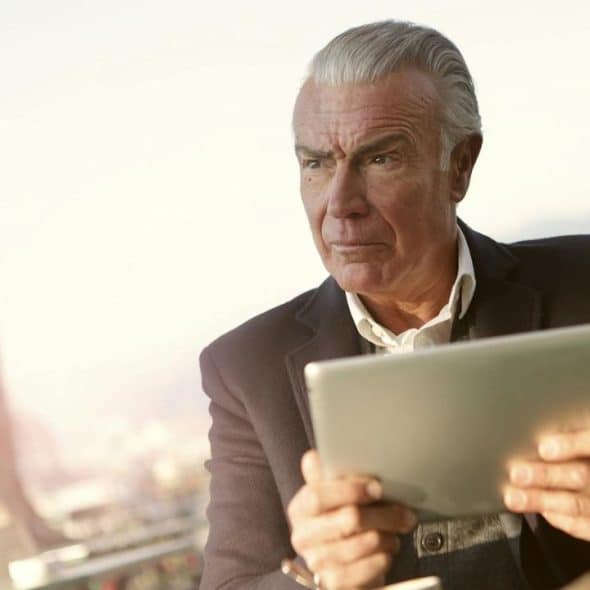A man with grey hair holds a tablet while looking away with a focused expression.