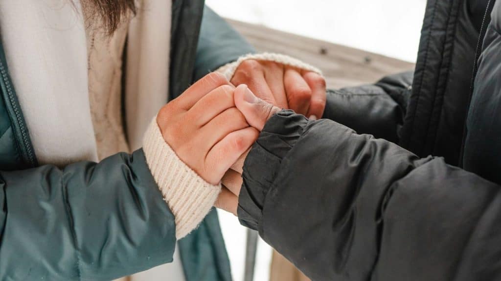 Close-up of two people in winter coats holding hands outdoors with a blurred background.
