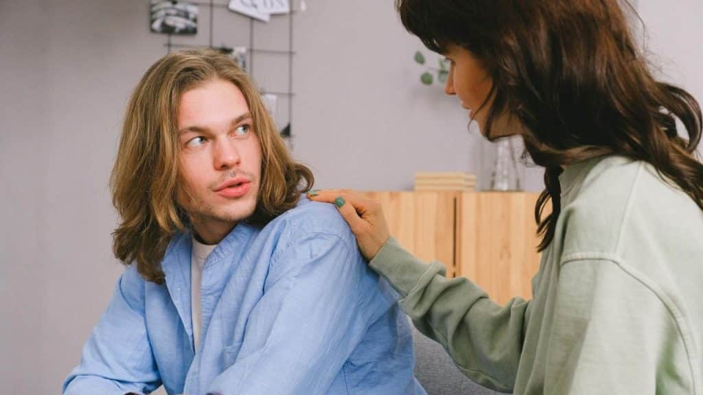 A person with long hair looks back at another person who has a hand resting on their shoulder.