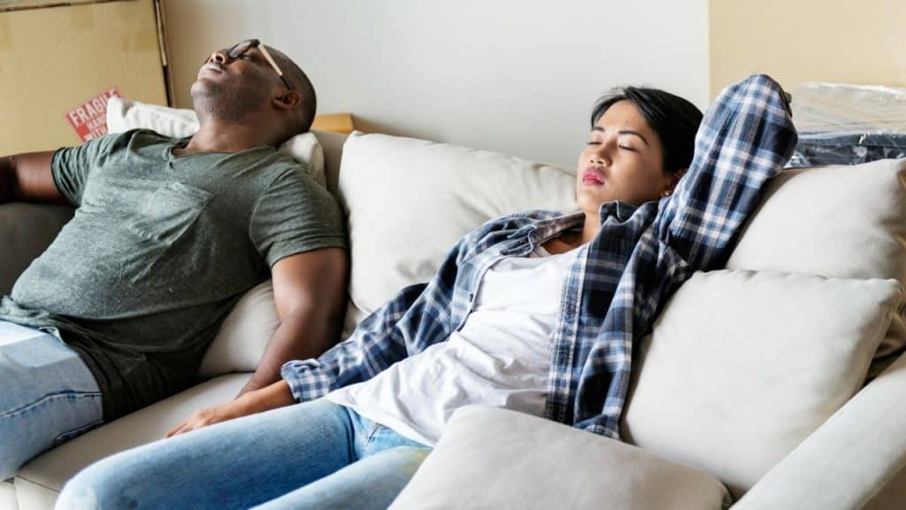 A man and woman resting on a light-colored sofa with their eyes closed indoors.