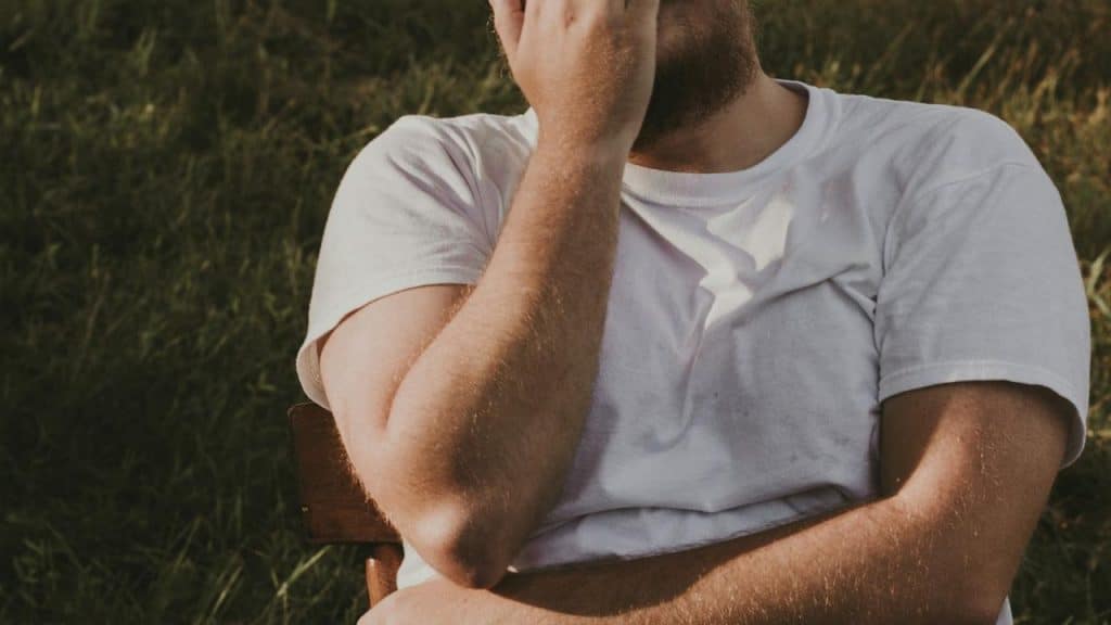 A man in a white t-shirt sitting outside with his hand covering part of his face.