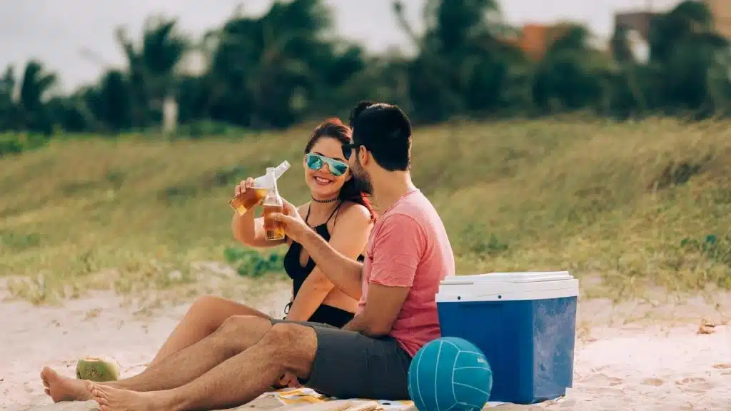A couple sitting on the beach clinking beer bottles.