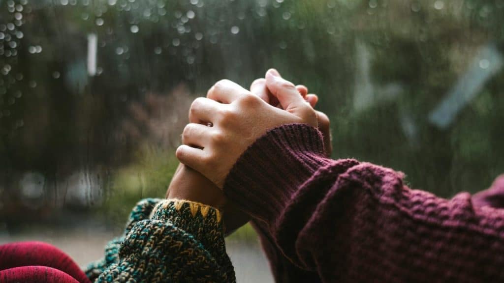 A close-up of two people holding hands by a rainy window.