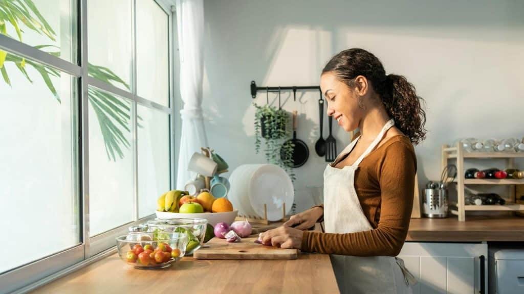 A woman in an apron chopping vegetables in a bright kitchen.