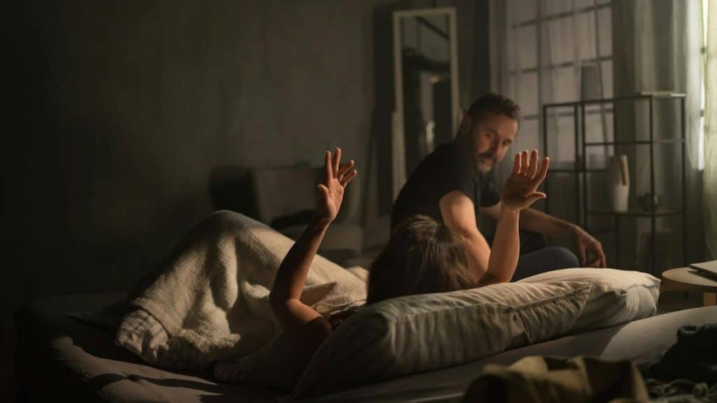 A person lying in bed raising their hands while someone sits nearby.