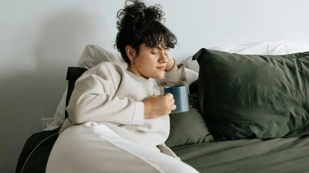 A woman reclining on a bed while holding a mug and resting her head against pillows.