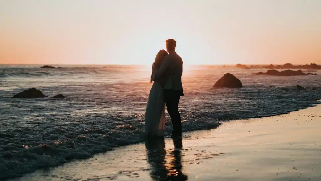 A couple standing close to each other while they stand on a beach and look at the sunset together.