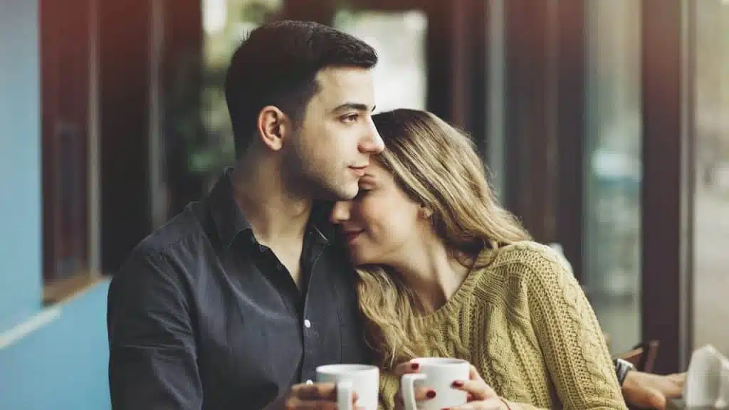 A woman smiling and snuggling with a man while they have coffee together.