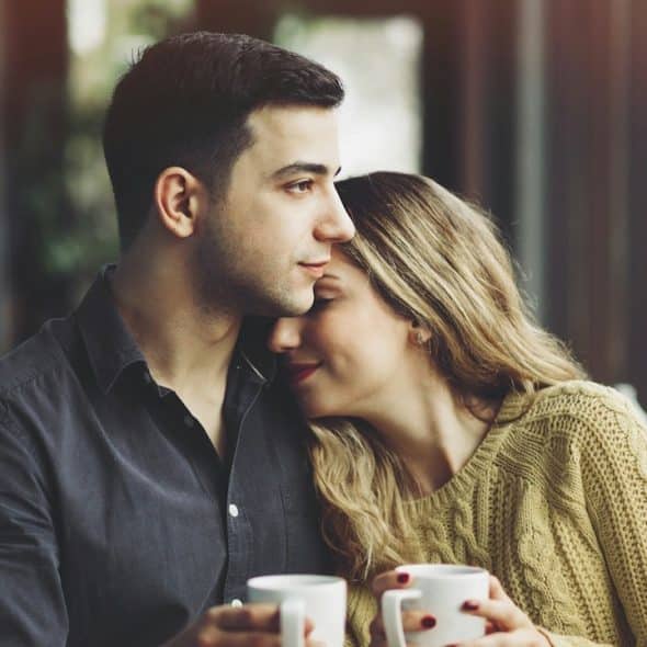 A woman smiling and snuggling with a man while they have coffee together.