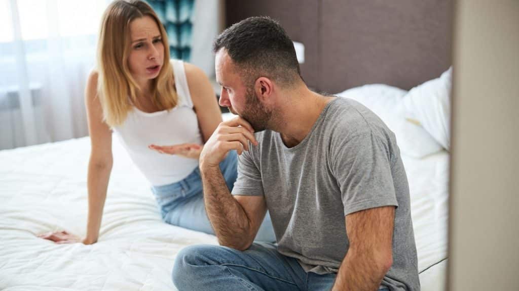 A woman talking to a man who’s struggling to listen to her while they sit on a bed.