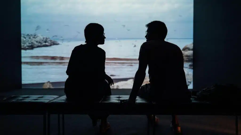 Silhouette of a couple sitting on a bench in front of a screen in a dimly lit room.