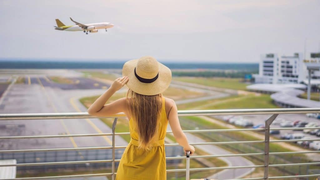 A woman wearing a hat adjusts her hat while looking at a plane taking off from a runway while she stands on a balcony overlooking it. 