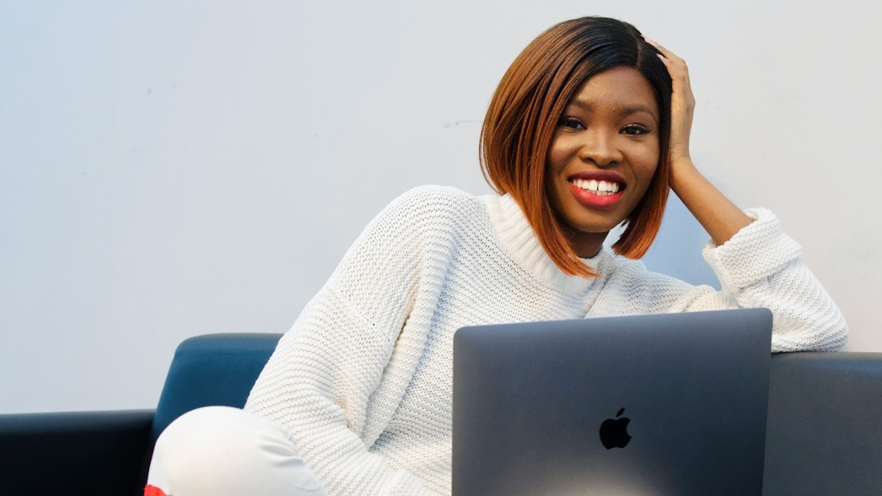 A woman using a laptop and smiling while sitting on a couch and supporting her head with her hand.
