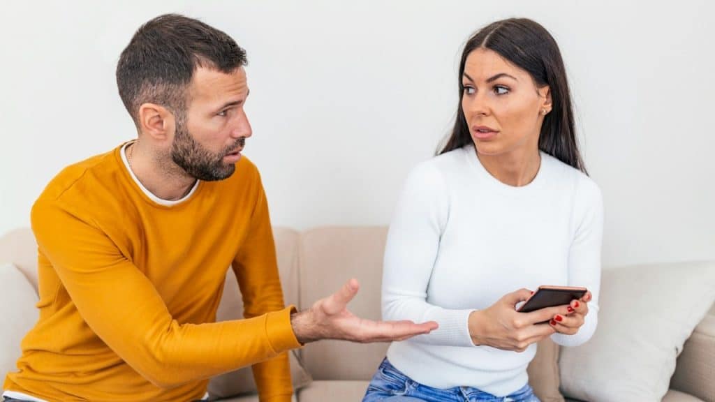 A woman looking at a man who’s asking for her phone as they sit on a couch.