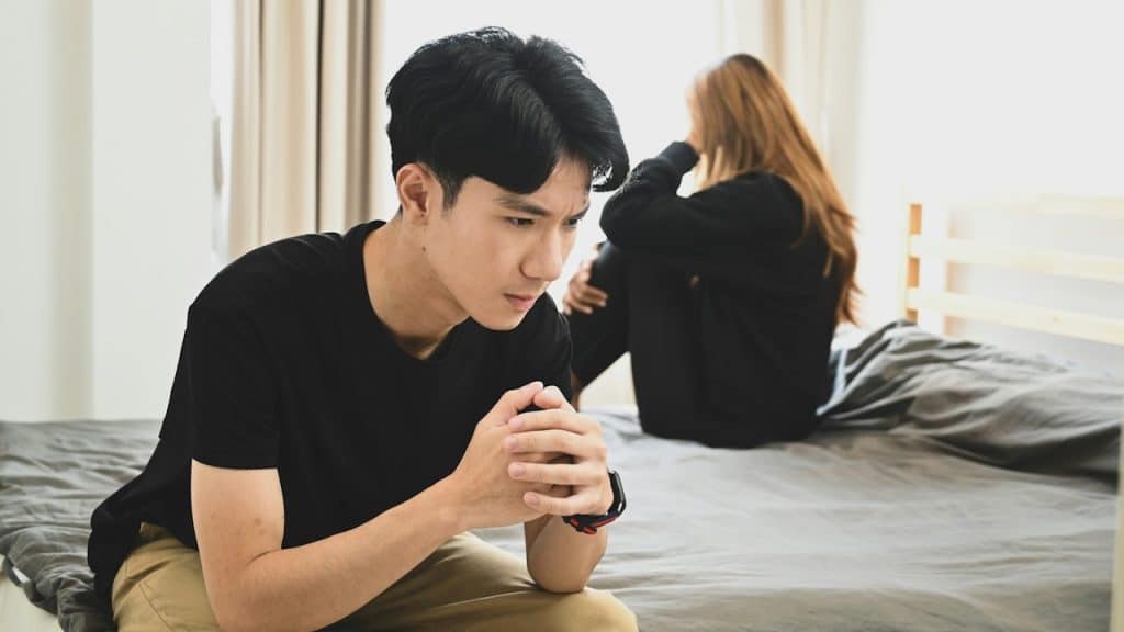 A man sitting on the edge of a bed looking distraught while a woman sits in the same bed behind him.
