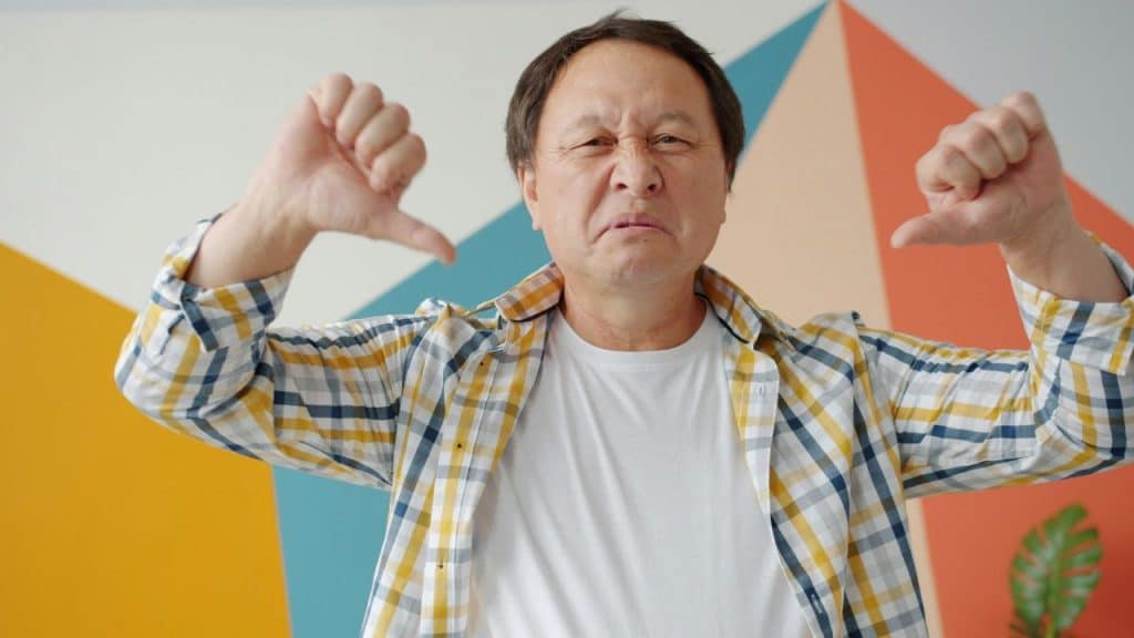 A middle-aged man in a plaid shirt gives a double thumbs-down with a grimace.