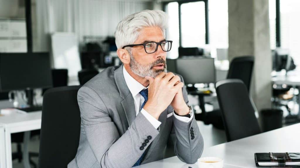 A man with grey hair and glasses sits at a desk with hands clasped.